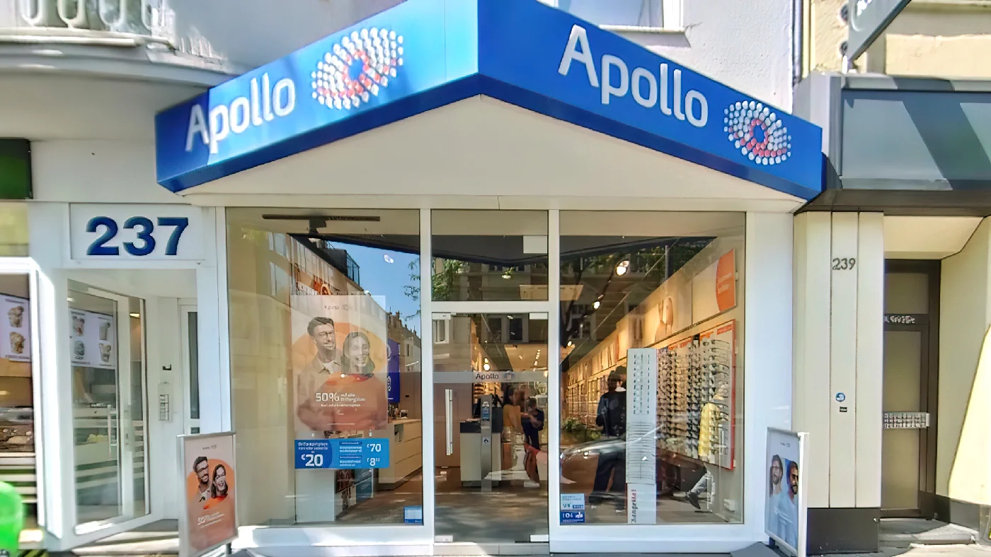Apollo-Optik Geschäft in Köln mit blauem Vordach weißem Logo Schaufenstern und Produkten an Straße mit Bäumen und Gebäuden. Augenoptiker Köln Nippes