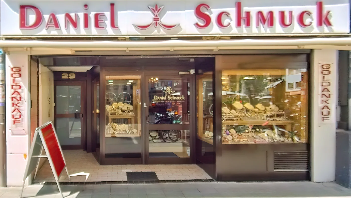 Geschäft Daniel Schmuck Köln rotes Logo Schild gut sichtbar zwei große Schaufenster voll Schmuck gläserne Eingangstür mit Name und Logo Sandwichschild auf Gehweg vor Laden Juwelier Köln Apostel-Viertel