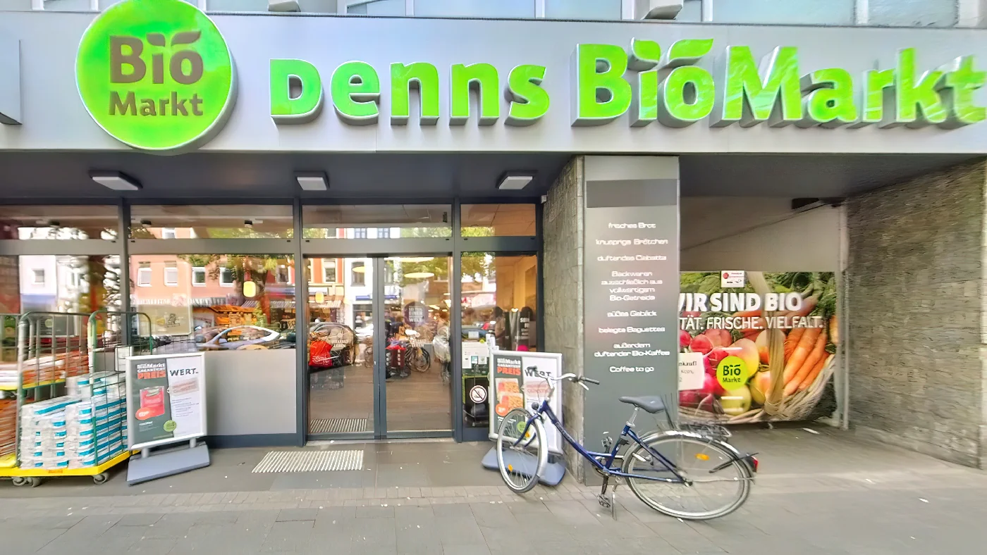 Denns BioMarkt grüner Schriftzug auf schwarzem Vordach zwei Fahrräder vorm Eingang Glastüren Werbeposter für Bio-Produkte in Fenstern Straße mit Bäumen und Gebäuden Bioladen Köln Lindenthal