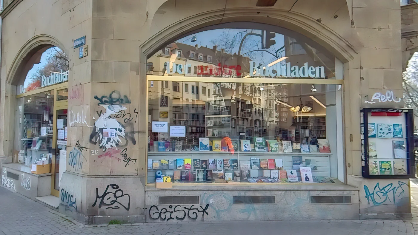Buchladen Der andere Buchladen in Steinbau mit Torbogen großes Schaufenster mit Büchern tagsüber geöffnet Straße mit Bürgersteig und Bäumen Graffiti auf Fassade Buchhandlung Köln Südstadt