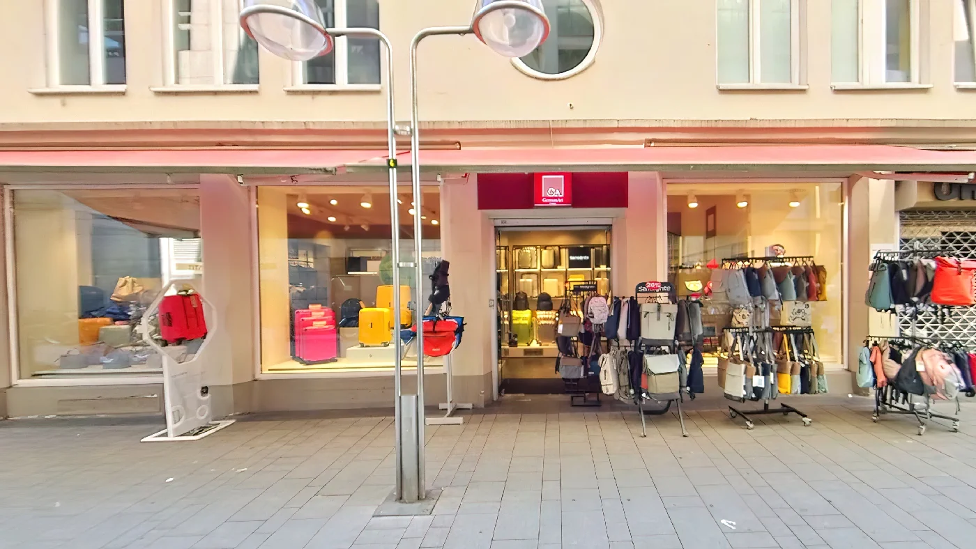 GA GermanArt in Köln mit roter C&A Markise Schaufenstern mit Kleidung und Accessoires Kleiderständern auf dem Bürgersteig zwei Straßenlaternen vor beiger Fassade bei blauem Himmel. Taschengeschäft Köln City