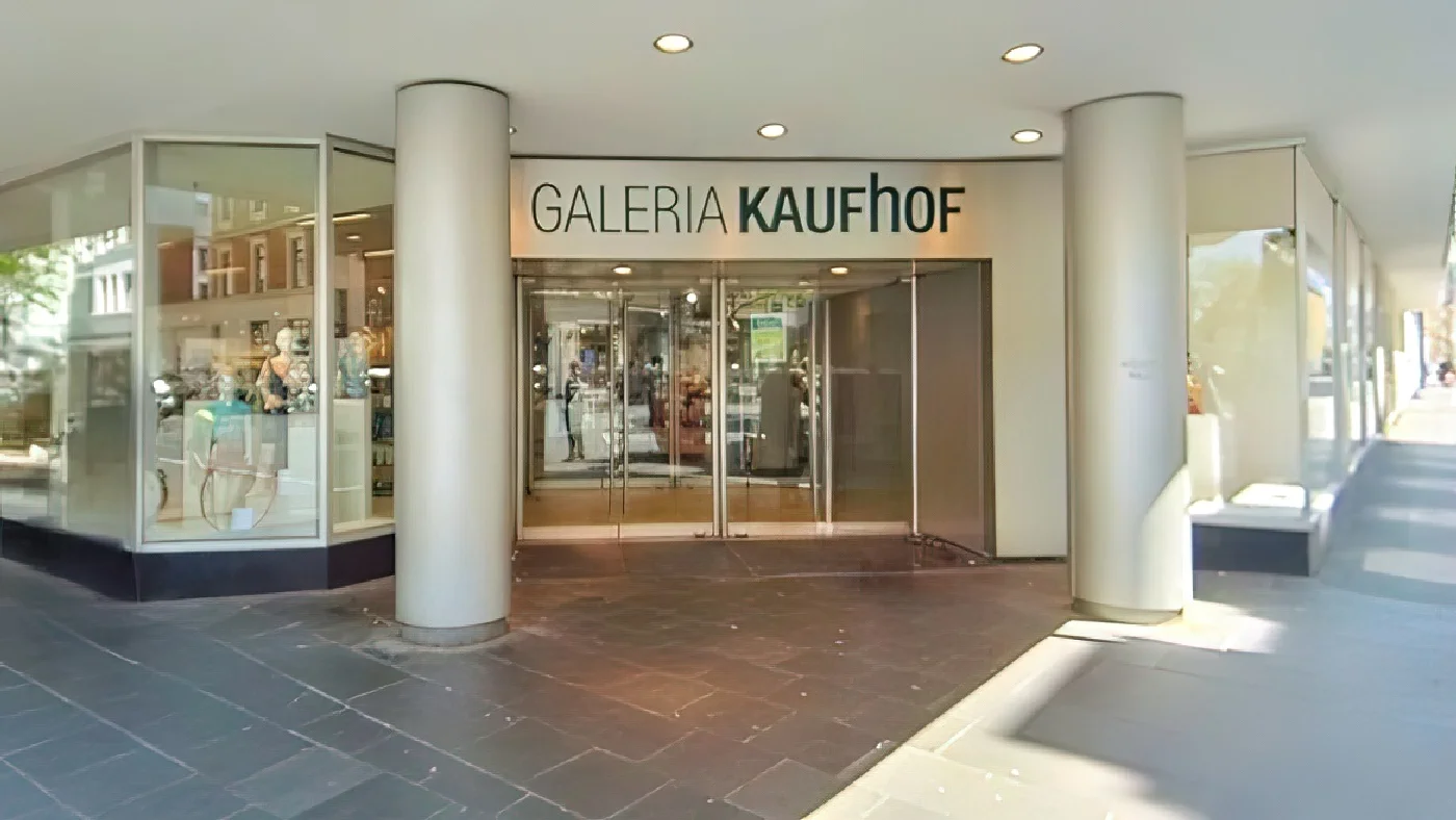GALERIA Köln-Nippes mit schwarzem Namen auf weißem Grund über Eingang zwei großen Glastüren mit Metallrahmen Schaufenstern mit Puppen und Kleidung an Straße mit Bäumen. Kaufhaus Köln Nippes